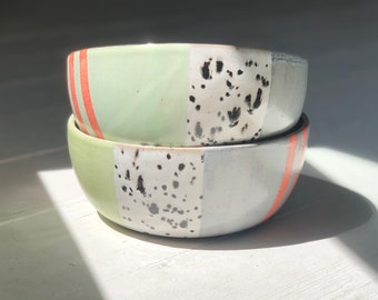 May include: Two ceramic bowls stacked on top of each other. The top bowl is light green with a white and black speckled design and a red stripe around the rim. The bottom bowl is white with a light green and black speckled design and a red stripe around the rim.