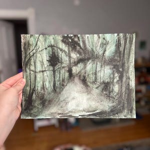 May include: A watercolor painting depicting a forest scene with a path leading into the distance. The artwork features a palette of dark greens, blacks, and whites, creating a moody and atmospheric landscape. The painting is held by a hand.