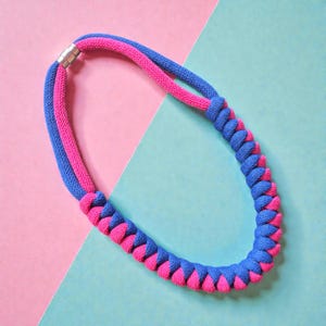May include: A striking necklace in a braided design, combining bright pink and blue. The necklace, with a magnetic clasp, is set against a pink and blue backdrop. This colourful accessory is a statement piece.