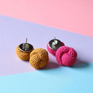May include: Close-up of two pairs of handmade stud earrings. One pair is mustard yellow, the other is hot pink. Each earring features a knotted, textured design and a silver-coloured post and backing.