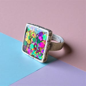 May include: A silver ring with a square face containing colourful glitter. The glitter features pink, green, yellow, and purple hues. The ring has a textured bezel and a plain band. The background is a mix of pink, blue, and purple.