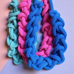 May include: Close-up of three colourful, braided rope necklaces. One is turquoise, one is pink, and one is blue. The necklaces are knotted and appear to be made of a thick, soft material. The background is a mix of pink and purple.