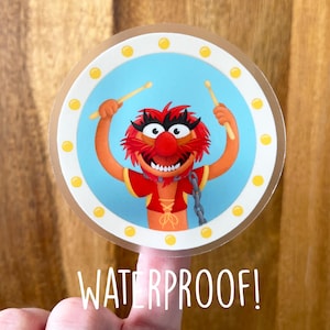 May include: A round sticker with a yellow border and a blue background. The sticker features a cartoon red and orange Muppet character playing drums with two drumsticks. The text "WATERPROOF!" is printed below the sticker.