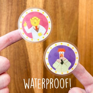 May include: Two round, clear, waterproof stickers with colorful images of characters from the Muppets. The first sticker features a yellow character with a white lab coat and a pink background. The second sticker features a red-haired character with a white lab coat and a purple background. The text "WATERPROOF!" is printed below the stickers.