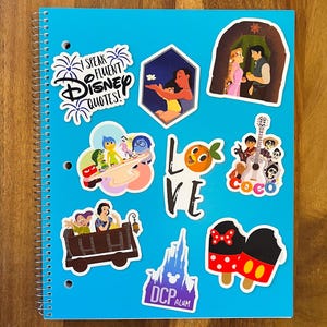 K&ouml;nnte beinhalten: Ein blaues Notizbuch mit verschiedenen Disney-Charakter-Stickern. Die Sticker zeigen Figuren aus Vaiana, Coco, Kopf hoch, Schneewittchen und Mickey Mouse. Das Notizbuch hat auch einen Sticker mit der Aufschrift "Ich spreche flie&szlig;end Disney-Zitate!"