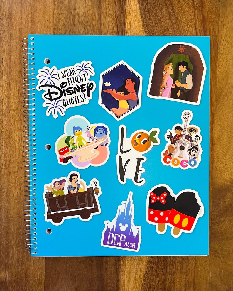 K&ouml;nnte beinhalten: Ein blaues Notizbuch mit verschiedenen Disney-Charakter-Stickern. Die Sticker zeigen Figuren aus Vaiana, Coco, Kopf hoch, Schneewittchen und Mickey Mouse. Das Notizbuch hat auch einen Sticker mit der Aufschrift "Ich spreche flie&szlig;end Disney-Zitate!"