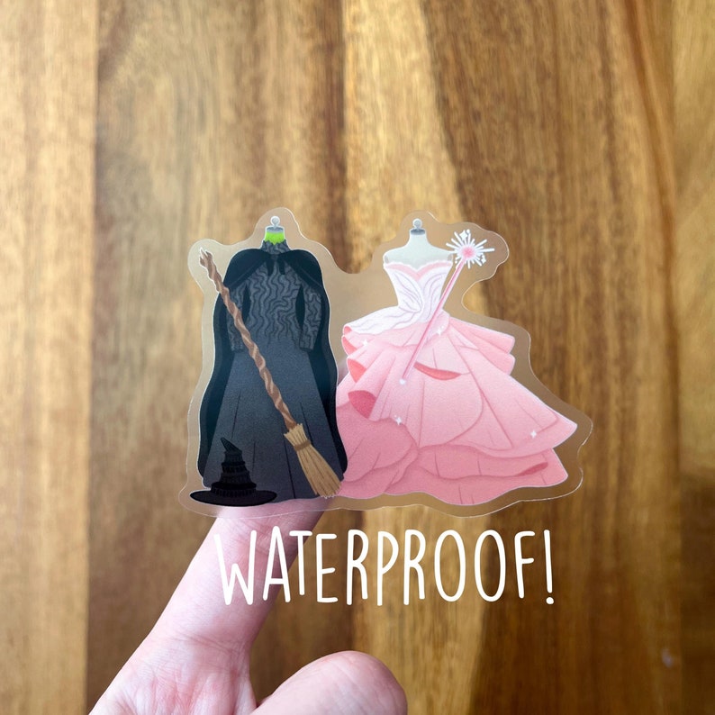 Puede incluir: Una pegatina transparente con una bruja negra y verde y un hada rosa y blanca. La pegatina dice "WATERPROOF!" en la parte inferior.