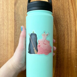 Puede incluir: Una botella de agua reutilizable azul claro con una pegatina de una bruja y una princesa. La pegatina es blanca con un contorno negro.