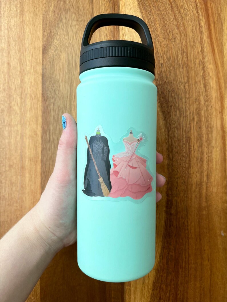 Puede incluir: Una botella de agua reutilizable azul claro con una pegatina de una bruja y una princesa. La pegatina es blanca con un contorno negro.