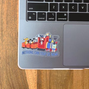 May include: A colorful sticker featuring a cartoon train with Mickey Mouse, Donald Duck, and Goofy. The train is red, blue, and yellow, with the text "NOTHING CAN STOP US NOW!" on the bottom. The sticker is on a laptop.