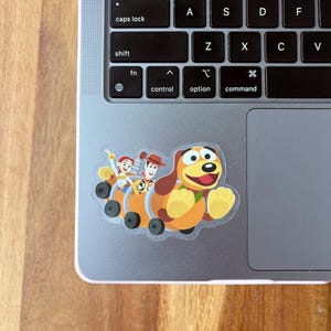 Puede incluir: Una pegatina transparente con una imagen de dibujos animados de Woody, Buzz Lightyear y Slinky Dog de la franquicia Toy Story. Los personajes van montados en el resorte de Slinky Dog.