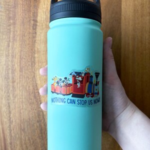 May include: A teal water bottle with a black handle and lid. A colorful sticker featuring cartoon characters on a train with the text "NOTHING CAN STOP US NOW!" is affixed to the bottle.