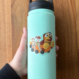 Puede incluir: Una botella de agua turquesa con tapa y asa negras. Una pegatina de Woody, Buzz Lightyear y Slinky Dog de Toy Story est&aacute; en la botella.