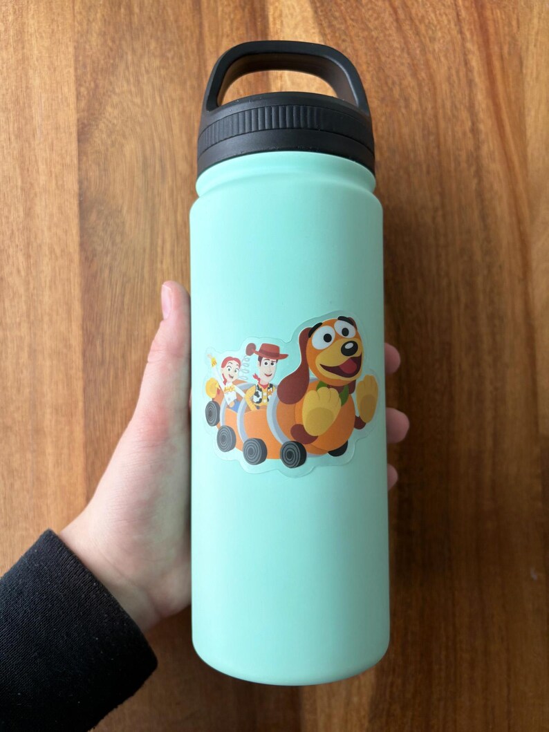 Puede incluir: Una botella de agua turquesa con tapa y asa negras. Una pegatina de Woody, Buzz Lightyear y Slinky Dog de Toy Story est&aacute; en la botella.