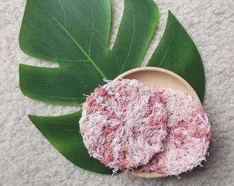 Puede incluir: Dos estropajos de crochet rosa y blanco en un plato de madera. Los estropajos están hechos de fibras naturales y son perfectos para exfoliar y limpiar la piel.