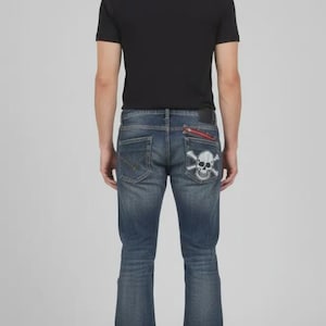 Puede incluir: Vaqueros de mezclilla de lavado oscuro con pernera acampanada. El bolsillo trasero presenta un dise&ntilde;o de calavera y huesos cruzados. Los vaqueros tienen un detalle de cremallera roja en el bolsillo trasero y una camiseta negra.