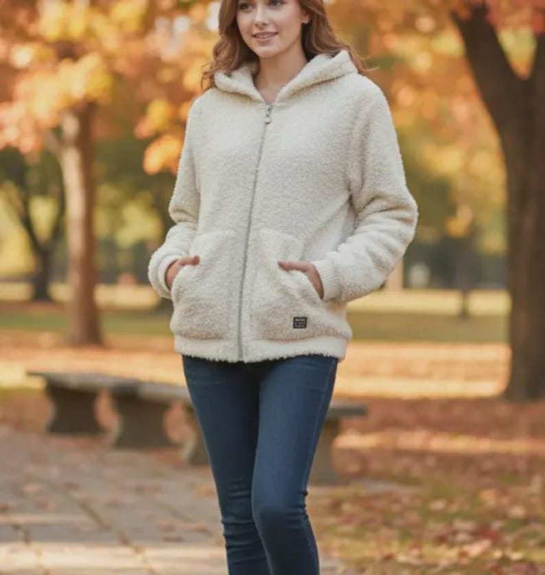 May include: Cream-colored hooded jacket with a full zipper closure and two front pockets. The jacket has a soft, textured appearance and a small square patch near the bottom. The person is wearing blue jeans. The background features a park setting with trees and a bench.
