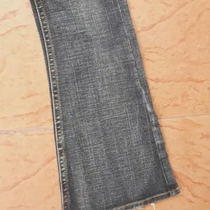 Puede incluir: Primer plano de un par de vaqueros de mezclilla azul descolorido. Los vaqueros tienen un ligero ensanchamiento en la parte inferior y presentan costuras visibles a lo largo de las costuras. El denim tiene un aspecto desgastado, con una decoloraci&oacute;n sutil y una superficie texturizada.