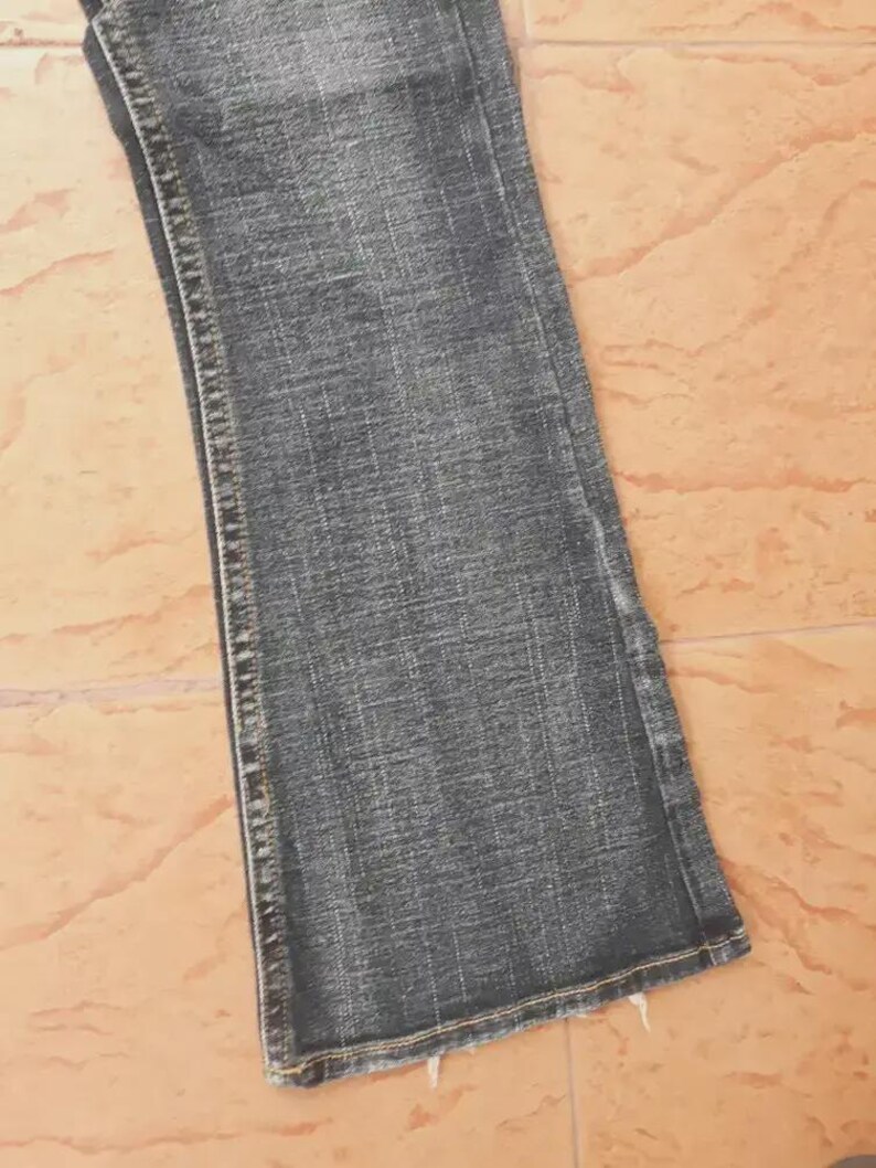 Puede incluir: Primer plano de un par de vaqueros de mezclilla azul descolorido. Los vaqueros tienen un ligero ensanchamiento en la parte inferior y presentan costuras visibles a lo largo de las costuras. El denim tiene un aspecto desgastado, con una decoloraci&oacute;n sutil y una superficie texturizada.