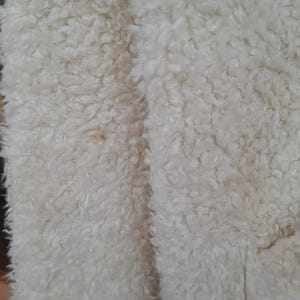 May include: Close-up of a cream-colored, textured fabric with a dense, curly pile. The material has a soft, plush appearance, with a slight variation in color. The fabric's surface is uneven, creating a tactile and visually interesting texture.