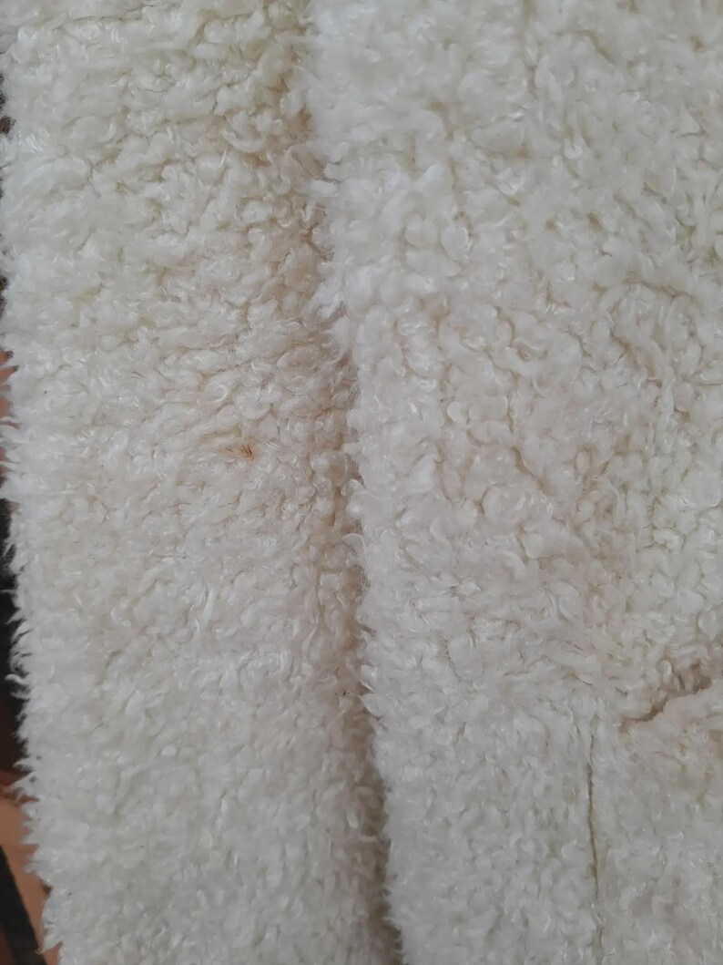 May include: Close-up of a cream-colored, textured fabric with a dense, curly pile. The material has a soft, plush appearance, with a slight variation in color. The fabric's surface is uneven, creating a tactile and visually interesting texture.