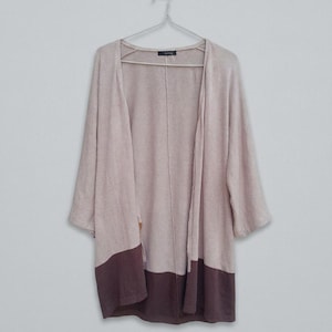 K&ouml;nnte beinhalten: Langer, offener Cardigan in einem weichen, ged&auml;mpften Rosa mit Dreiviertel&auml;rmeln. Der untere Teil des Cardigans ist in einem dunkleren Braunton gehalten. Der Cardigan h&auml;ngt an einem Metallb&uuml;gel vor einem schlichten wei&szlig;en Hintergrund.