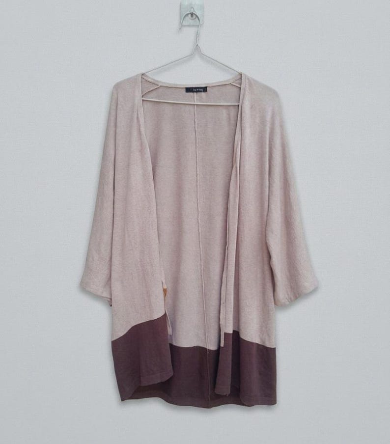 K&ouml;nnte beinhalten: Langer, offener Cardigan in einem weichen, ged&auml;mpften Rosa mit Dreiviertel&auml;rmeln. Der untere Teil des Cardigans ist in einem dunkleren Braunton gehalten. Der Cardigan h&auml;ngt an einem Metallb&uuml;gel vor einem schlichten wei&szlig;en Hintergrund.