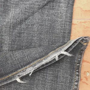 Puede incluir: Primer plano de unos vaqueros de mezclilla gris oscuro. La tela tiene un aspecto texturizado con l&iacute;neas de tejido verticales y horizontales visibles. El dobladillo est&aacute; deshilachado y las costuras son visibles a lo largo de las costuras.