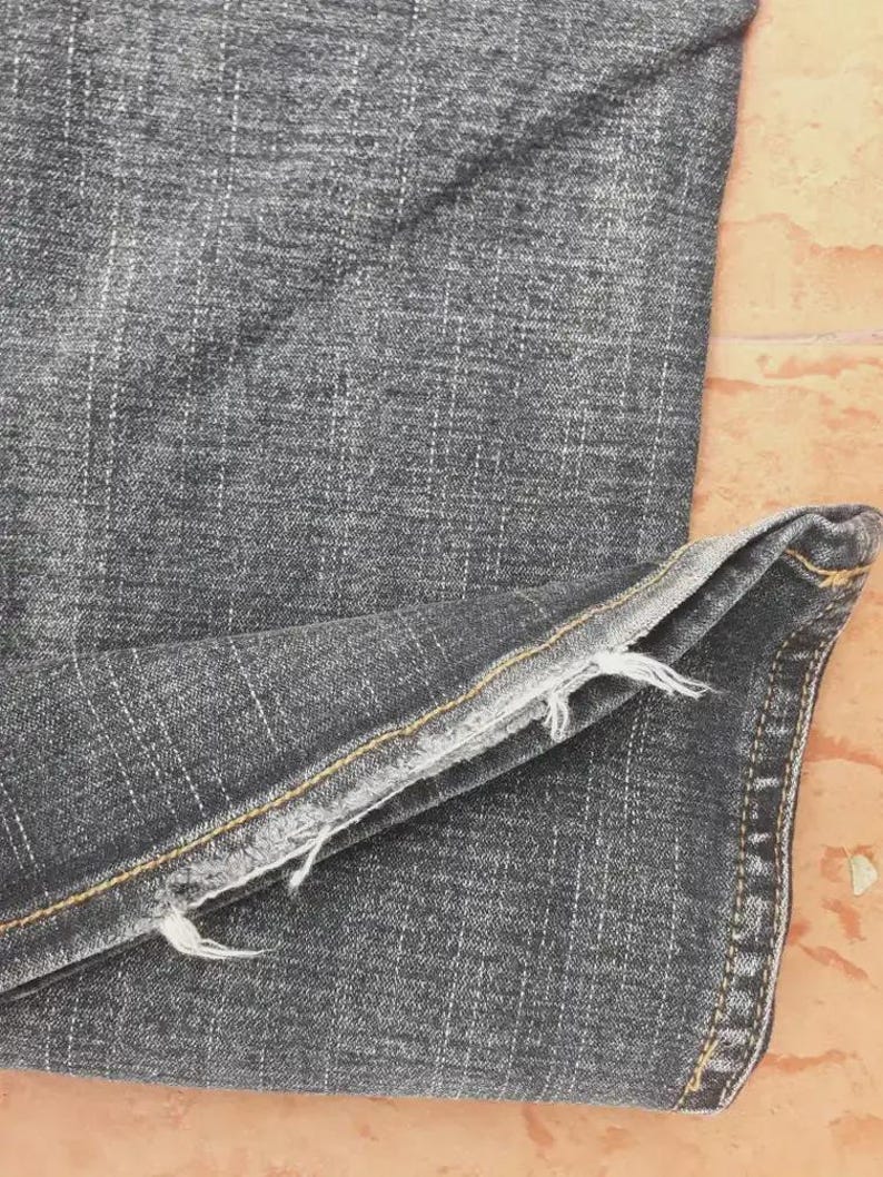 Puede incluir: Primer plano de unos vaqueros de mezclilla gris oscuro. La tela tiene un aspecto texturizado con l&iacute;neas de tejido verticales y horizontales visibles. El dobladillo est&aacute; deshilachado y las costuras son visibles a lo largo de las costuras.