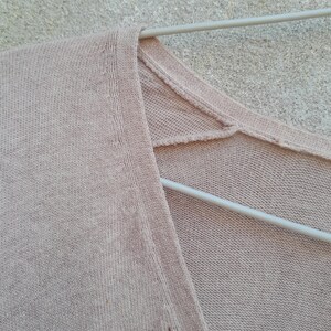 K&ouml;nnte beinhalten: Nahaufnahme eines hellrosa V-Ausschnitt-Sweaters, der an einem wei&szlig;en Kleiderb&uuml;gel h&auml;ngt. Der Strickstoff hat eine subtile Textur und eine weiche, ged&auml;mpfte Farbe. Der Halsausschnitt und die Schultern&auml;hte sind sichtbar.