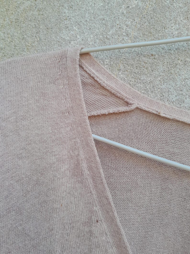 K&ouml;nnte beinhalten: Nahaufnahme eines hellrosa V-Ausschnitt-Sweaters, der an einem wei&szlig;en Kleiderb&uuml;gel h&auml;ngt. Der Strickstoff hat eine subtile Textur und eine weiche, ged&auml;mpfte Farbe. Der Halsausschnitt und die Schultern&auml;hte sind sichtbar.