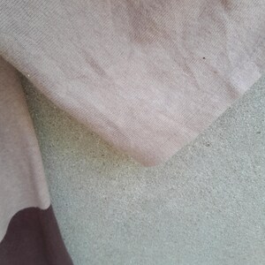 K&ouml;nnte beinhalten: Nahaufnahme eines beigen und braunen Stoffes. Der Stoff scheint ein weiches Strickmaterial zu sein, mit einem dunkleren braunen Abschnitt am unteren Rand. Der Hintergrund ist eine strukturierte, hellgraue Oberfl&auml;che.
