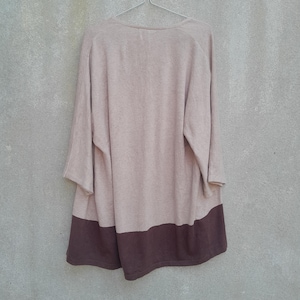 K&ouml;nnte beinhalten: Ein lang&auml;rmliger, beige-brauner Farbblock-Pullover h&auml;ngt an einem Metallb&uuml;gel vor einer grauen Wand. Der Pullover hat einen weiten Ausschnitt und dreiviertellange &Auml;rmel. Der untere Teil des Pullovers ist dunkelbraun.