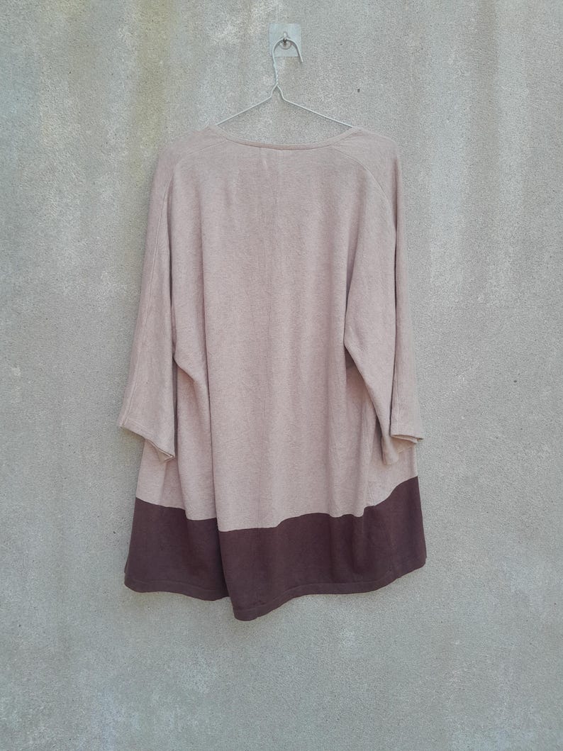 K&ouml;nnte beinhalten: Ein lang&auml;rmliger, beige-brauner Farbblock-Pullover h&auml;ngt an einem Metallb&uuml;gel vor einer grauen Wand. Der Pullover hat einen weiten Ausschnitt und dreiviertellange &Auml;rmel. Der untere Teil des Pullovers ist dunkelbraun.