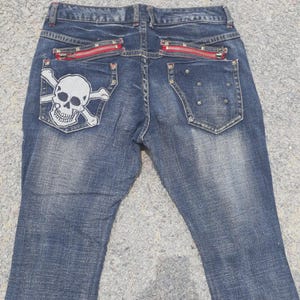 Puede incluir: Vaqueros azules con un parche de calavera y huesos cruzados en el bolsillo trasero. Los vaqueros tienen detalles de cremalleras rojas en los bolsillos traseros. La mezclilla tiene un lavado descolorido. Estilo casual.