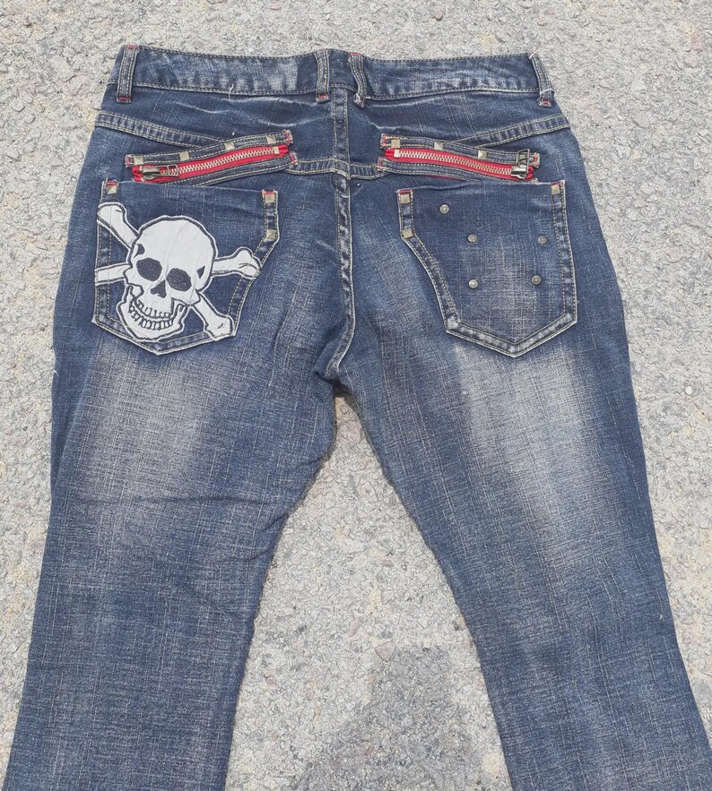 Puede incluir: Vaqueros azules con un parche de calavera y huesos cruzados en el bolsillo trasero. Los vaqueros tienen detalles de cremalleras rojas en los bolsillos traseros. La mezclilla tiene un lavado descolorido. Estilo casual.