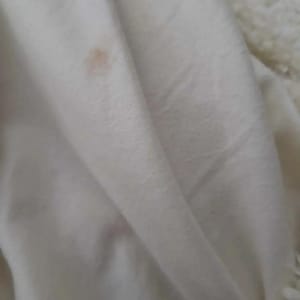 May include: Close-up of a white, folded blanket with a small, light brown stain. The blanket appears to be made of a soft, plush material, possibly fleece or a similar fabric. The texture is visible in the folds.