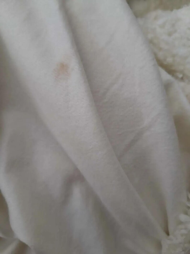 May include: Close-up of a white, folded blanket with a small, light brown stain. The blanket appears to be made of a soft, plush material, possibly fleece or a similar fabric. The texture is visible in the folds.