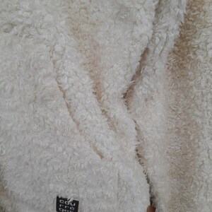 May include: Close-up of a cream-colored, fluffy jacket. The jacket has a textured, shaggy appearance. A small black square tag with white text is visible on the lower left side. The jacket appears soft and warm, suitable for cold weather.
