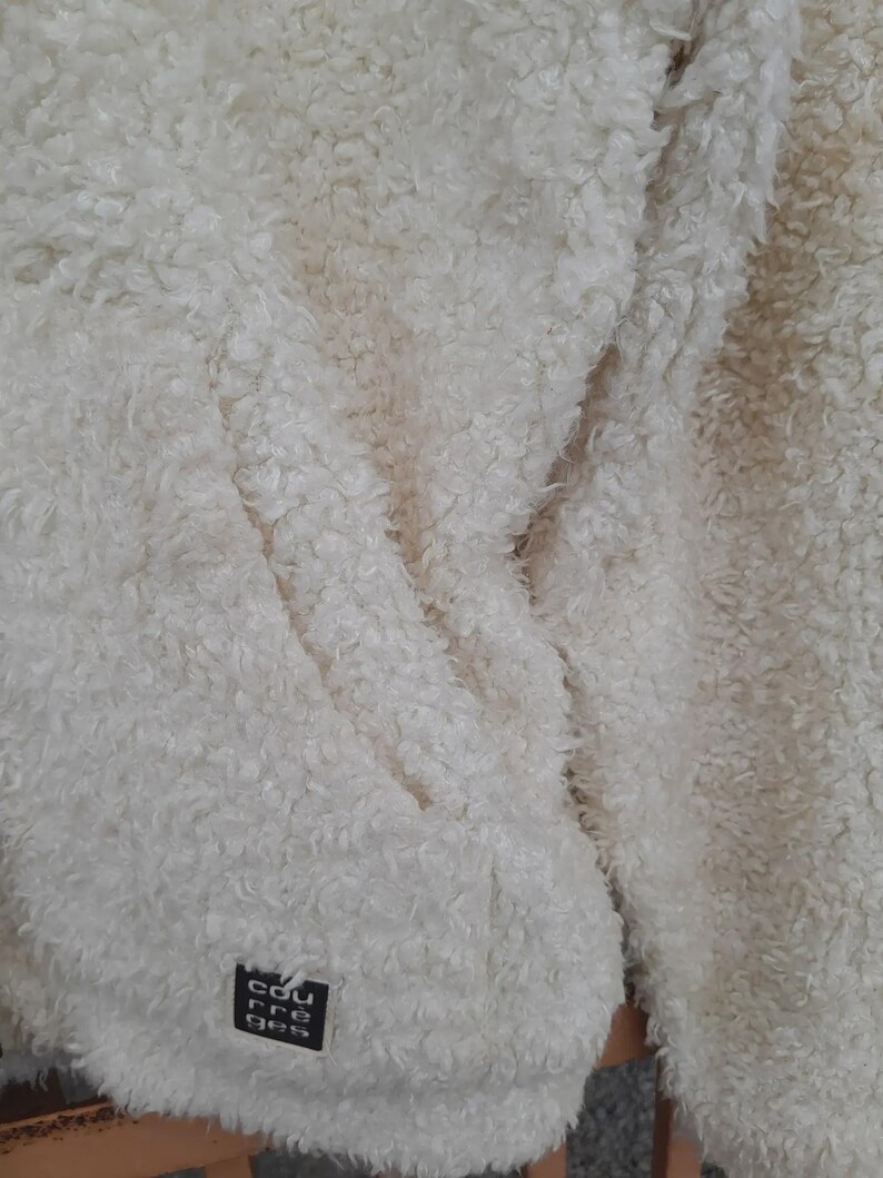 May include: Close-up of a cream-colored, fluffy jacket. The jacket has a textured, shaggy appearance. A small black square tag with white text is visible on the lower left side. The jacket appears soft and warm, suitable for cold weather.