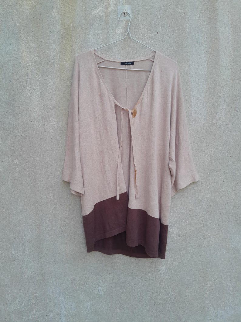 K&ouml;nnte beinhalten: Ein beige-brauner Cardigan mit Farbblock-Design, V-Ausschnitt und Dreiviertel&auml;rmeln h&auml;ngt an einem Metallb&uuml;gel. Der obere Teil ist beige, der untere Teil ist dunkelbraun. Der Cardigan hat einen Bindverschluss und Holzkn&ouml;pfe.