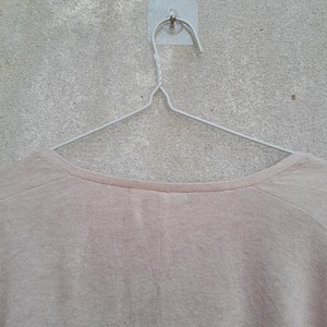 K&ouml;nnte beinhalten: Ein hellbeige Rundhals-Shirt h&auml;ngt an einem silbernen Metallb&uuml;gel. Das Shirt besteht aus einem weichen, strukturierten Stoff. Der Kleiderb&uuml;gel ist mit einem kleinen, quadratischen Klebehaken an einer grauen Wand befestigt.