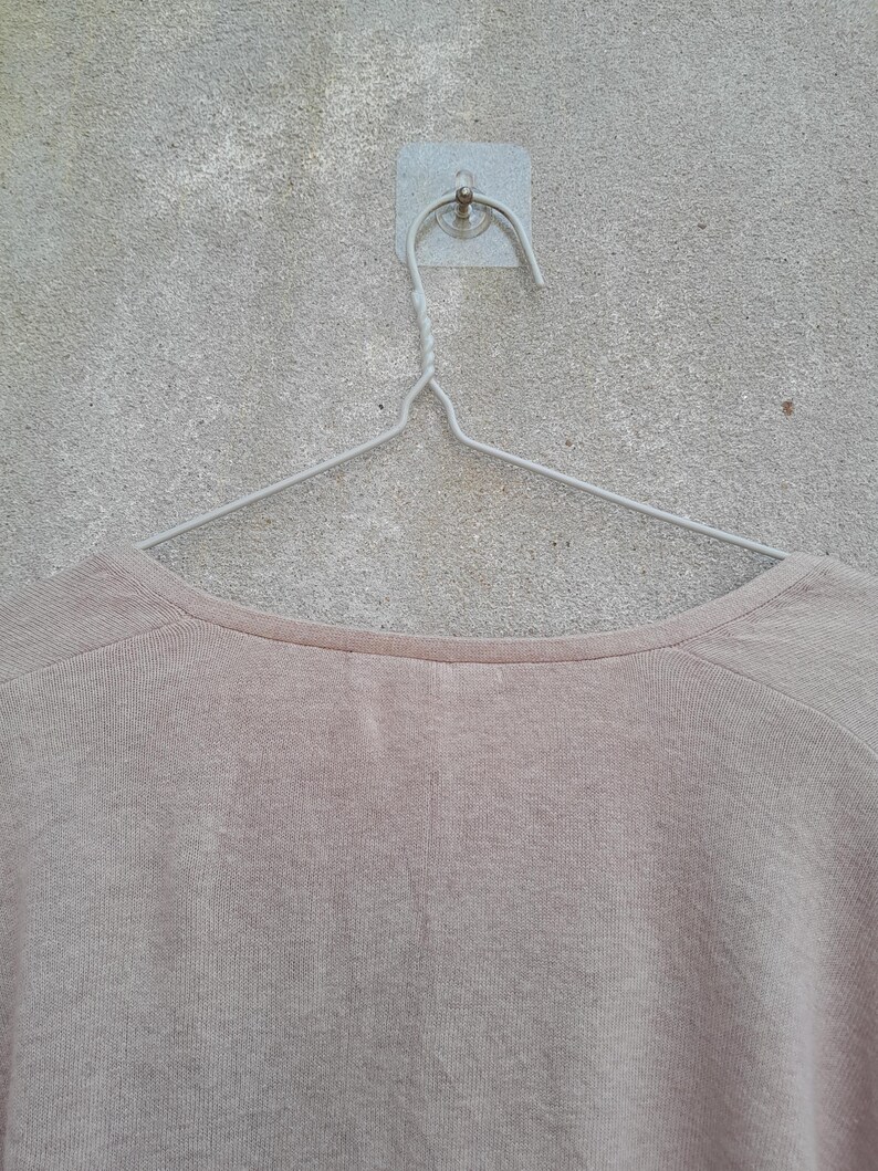 K&ouml;nnte beinhalten: Ein hellbeige Rundhals-Shirt h&auml;ngt an einem silbernen Metallb&uuml;gel. Das Shirt besteht aus einem weichen, strukturierten Stoff. Der Kleiderb&uuml;gel ist mit einem kleinen, quadratischen Klebehaken an einer grauen Wand befestigt.