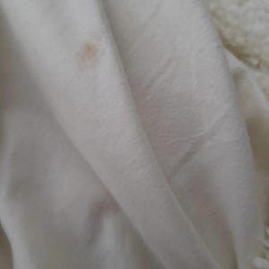 May include: Close-up of a white, folded fabric with a small, light brown stain. The fabric appears soft and possibly made of a plush material. The texture is visible in the folds and shadows.
