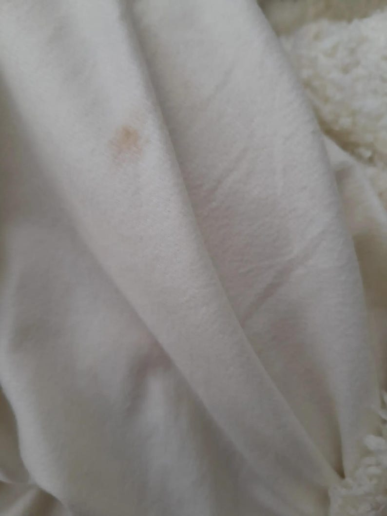 May include: Close-up of a white, folded fabric with a small, light brown stain. The fabric appears soft and possibly made of a plush material. The texture is visible in the folds and shadows.