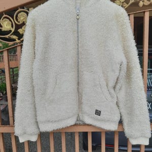 May include: Cream-colored, hooded fleece jacket with a full zipper closure. The jacket has a soft, textured appearance, ribbed cuffs, and a small square patch near the bottom. The jacket is hanging on a metal fence.