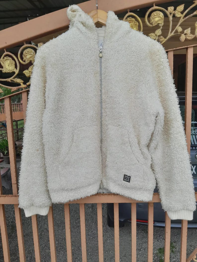 May include: Cream-colored, hooded fleece jacket with a full zipper closure. The jacket has a soft, textured appearance, ribbed cuffs, and a small square patch near the bottom. The jacket is hanging on a metal fence.