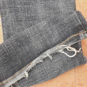 Puede incluir: Primer plano de unos vaqueros de mezclilla gris oscuro. Los vaqueros est&aacute;n doblados, revelando el dobladillo deshilachado y las costuras. La tela tiene un aspecto texturizado con patrones de tejido verticales y horizontales visibles. El aspecto general es informal y desgastado.