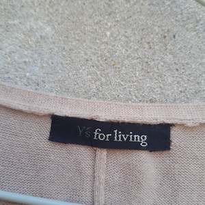 K&ouml;nnte beinhalten: Nahaufnahme eines beigefarbenen Strickkleidungsst&uuml;cks mit einem dunkelblauen Etikett. Auf dem Etikett steht "Y's for living" in wei&szlig;er Schrift. Das Kleidungsst&uuml;ck h&auml;ngt an einem wei&szlig;en Kleiderb&uuml;gel vor einem strukturierten Hintergrund.