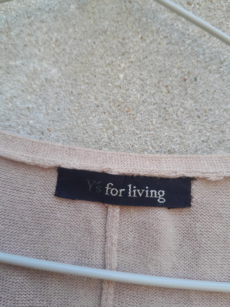 K&ouml;nnte beinhalten: Nahaufnahme eines beigefarbenen Strickkleidungsst&uuml;cks mit einem dunkelblauen Etikett. Auf dem Etikett steht "Y's for living" in wei&szlig;er Schrift. Das Kleidungsst&uuml;ck h&auml;ngt an einem wei&szlig;en Kleiderb&uuml;gel vor einem strukturierten Hintergrund.
